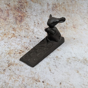 May include: A black cast iron doorstop shaped like a mouse holding a small ball. The doorstop has a textured surface with a pattern of circles.
