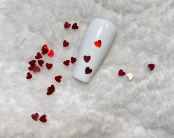 May include: White fake nail with red glitter hearts on a white fuzzy surface. There are also red glitter hearts scattered around the nail.