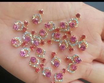 May include: Assortment of pink and clear rhinestone embellishments with gold settings. The rhinestones are in various shapes and sizes, including round, square, and teardrop.