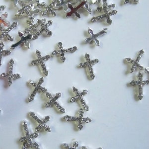 May include: A scattering of silver-toned cross-shaped charms, each adorned with small, clear rhinestones. The charms are scattered on a white surface.
