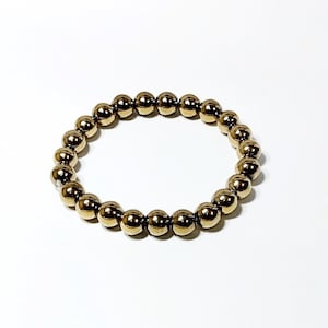 Puede incluir: Pulsera de cuentas doradas. La pulsera está hecha de cuentas redondas y brillantes ensartadas. Las cuentas tienen un brillo metálico y reflejan la luz. La pulsera se muestra sobre un fondo blanco.