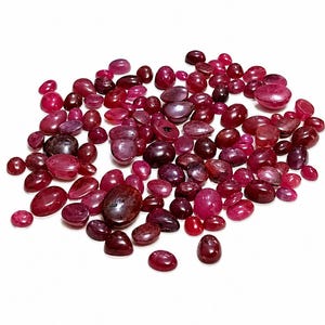 Puede incluir: Una colección de piedras preciosas de rubí pulidas en varias formas y tamaños. Las piedras preciosas varían en color, desde el rojo intenso hasta el rojo púrpura, con una superficie brillante y reflectante. Las gemas están dispersas sobre un fondo blanco.