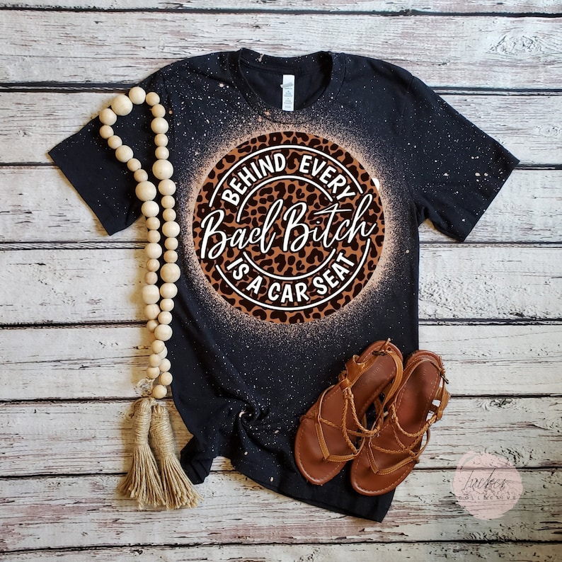 Puede incluir: Camiseta negra con dise&ntilde;o blanqueado y un c&iacute;rculo con estampado de leopardo. El c&iacute;rculo contiene el texto "BEHIND EVERY Bad Bitch IS A CAR SEAT" en una mezcla de fuentes. Sandalias marrones y un collar de cuentas tambi&eacute;n est&aacute;n en la imagen.