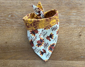 May include: A Thanksgiving-themed dog bandana. The bandana features a white background with cartoon turkeys and the text "Gobble 'Til You Wobble!" The reverse side has a sunflower pattern in shades of yellow and brown.