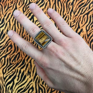 Könnte beinhalten: Ein silberner Ring mit einem großen, quadratischen Tigeraugenstein. Der Stein hat eine warme braune Farbe mit natürlichen Mustern. Der Ring hat ein gedrehtes Seildesign um den Stein, und das Band ist silber. Der Ring wird an einer Hand gezeigt.
