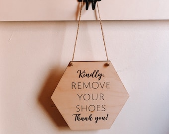 May include: A wooden hexagonal sign with the text "Kindly, Remove Your Shoes Thank you!" hanging from a black hook on a white wall.