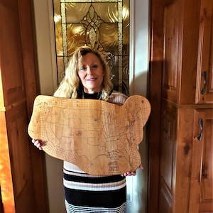 May include: A woman holds a wooden game board shaped like the United States. The board is a map of the United States with each state outlined. The board is called "Orts Mapa de States" and has the words "Adventure Boards" at the bottom.