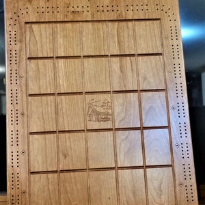 May include: Wooden cribbage board with the text "THE LARSON LODGE Things are Simpler at the Cabin" engraved at the top. The board has a grid of squares and holes for pegs, with a cabin illustration in the center. Numbers 1-5 are at the bottom.