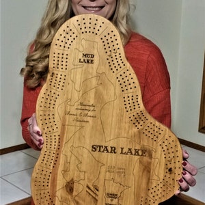 May include: A wooden game board shaped like the outline of a lake, with the text "MUD LAKE" and "STAR LAKE" printed on it. The board is decorated with holes and a small map of Minnesota. The text "Handcrafted exclusively for Bonnie & Ronnie Fricsanen" is also printed on the board.