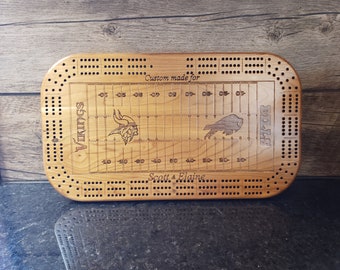 May include: A wooden cribbage board with a custom football field design. The board features the logos of the Minnesota Vikings and Buffalo Bills, along with the text "Custom made for Scott & Elaine".