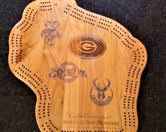 May include: A wooden cribbage board shaped like the state of Wisconsin, featuring logos for the Green Bay Packers, Milwaukee Brewers, and Milwaukee Bucks. The board is personalized with the text "Crafted exclusively for Marsha & Dale Thompson".