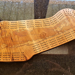 May include: A wooden cribbage board shaped like Long Island, New York. The board has a natural wood finish and features the names of various towns and cities on Long Island.