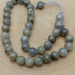 May include: A strand of round, gray-green gemstone beads, likely labradorite, with a subtle sheen. The beads are strung together, forming a loop, and are displayed on a wooden surface. The beads are approximately 0.8 cm in diameter.