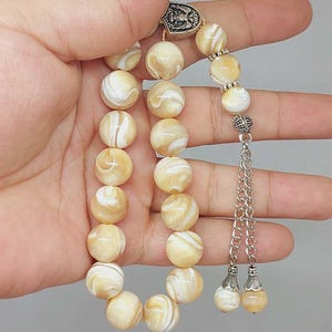 May include: A strand of prayer beads with creamy white and tan marbled beads. The beads are connected by a silver chain and decorative accents. The beads are held in a hand.