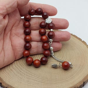 Op de afbeelding: Een gebedskralenrozenkrans met donkerroodbruine houten kralen. De ronde kralen zijn geregen met een zilverkleurige ketting en decoratieve elementen. De rozenkrans wordt getoond op een houten oppervlak.