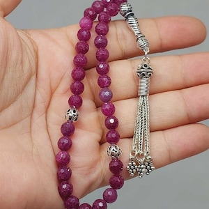 Puede incluir: Un rosario de cuentas de piedra preciosa de color rosa oscuro con detalles plateados y un borla. Las cuentas son redondas y tienen una textura facetada. La borla está hecha de cadenas plateadas y tiene un pequeño dije plateado al final.