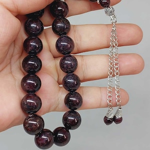 Puede incluir: Un collar de cuentas de oración de piedras preciosas de color rojo oscuro con detalles plateados y un borla. Las cuentas son redondas y tienen un acabado liso y pulido.