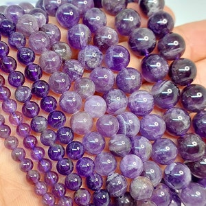 May include: Strands of polished amethyst beads in varying sizes, ranging from small to large. The beads display a gradient of purple hues, from deep violet to lighter shades. The round beads are strung together, showcasing their natural color and texture.