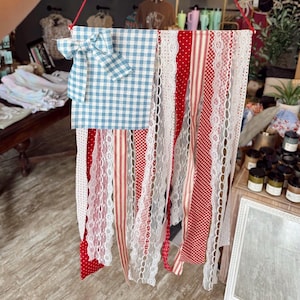 May include: A decorative wall hanging resembling an American flag. It features a blue and white gingham bow and a matching square. Red and white striped and dotted ribbons, along with white lace, form the flag's stripes.