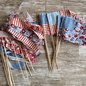 May include: A collection of fabric flags in red, white, and blue patterns, some with wooden sticks. The flags feature floral, striped, and dotted designs, perfect for patriotic or festive decorations. The flags are arranged on a wooden surface.