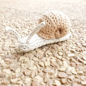 May include: A crocheted snail toy with a beige shell and a white body. The snail is sitting on a gravel surface.