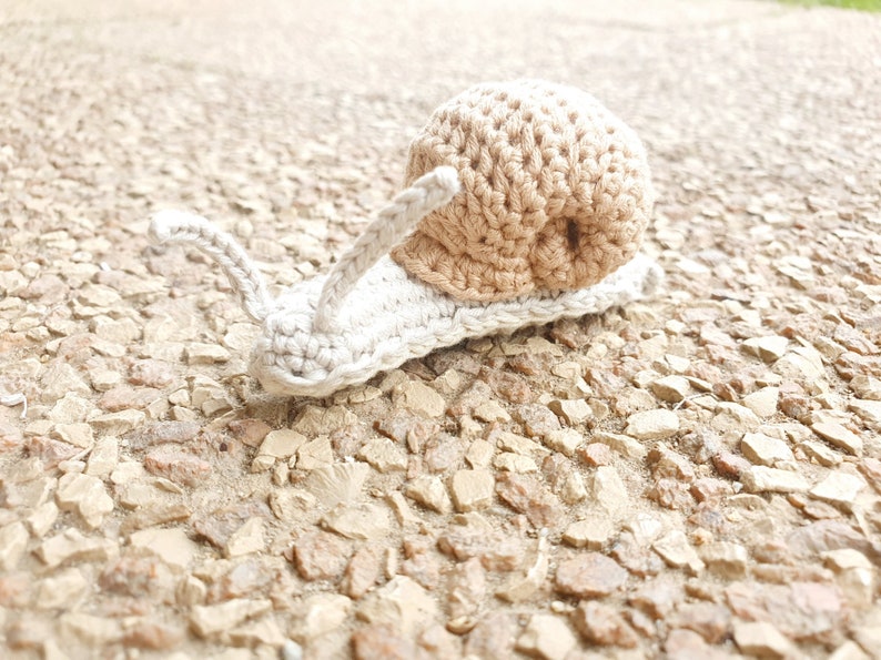 May include: A crocheted snail toy with a beige shell and a white body. The snail is sitting on a gravel surface.