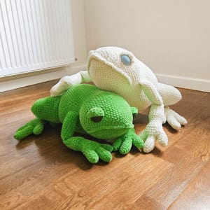Könnte beinhalten: Zwei handgefertigte Frosch-Plüschtiere aus Häkelarbeit. Eines ist hellgrün, das andere ist weiß. Der grüne Frosch liegt, während der weiße Frosch oben positioniert ist. Beide haben detaillierte Merkmale, einschließlich Augen und strukturierter Körper.