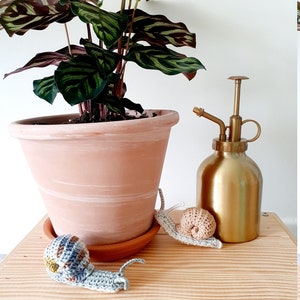 May include: Two crocheted snails, one gray and one beige, on a wooden surface next to a potted plant and a gold metal watering can.