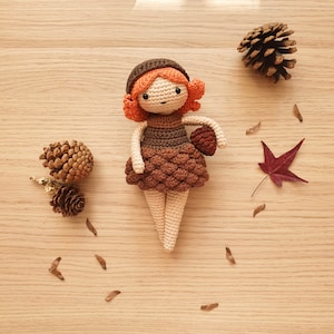 May include: A crocheted doll with brown hair, a brown dress, and a brown hat. The doll is holding a small brown acorn. The doll is sitting on a wooden surface with brown pine cones and a brown leaf.