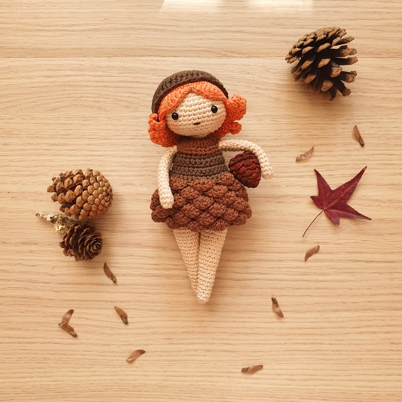 May include: A crocheted doll with brown hair, a brown dress, and a brown hat. The doll is holding a small brown acorn. The doll is sitting on a wooden surface with brown pine cones and a brown leaf.
