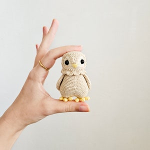 K&ouml;nnte beinhalten: Eine kleine geh&auml;kelte Eule mit gelben F&uuml;&szlig;en und schwarzen Augen. Die Eule ist beige und hat einen wei&szlig;en geh&auml;kelten Kragen.