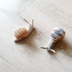 May include: Two crocheted snail toys. One snail is beige with a brown shell. The other snail is gray with a blue and brown striped shell.