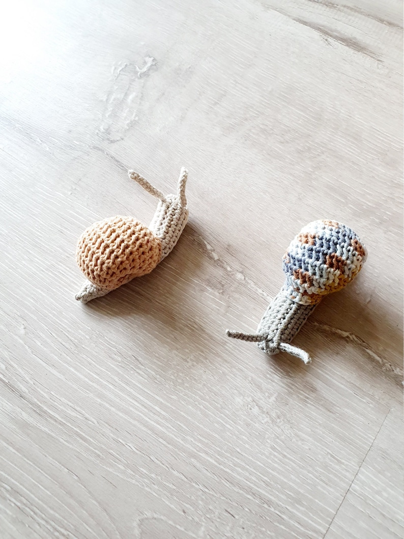 May include: Two crocheted snail toys. One snail is beige with a brown shell. The other snail is gray with a blue and brown striped shell.