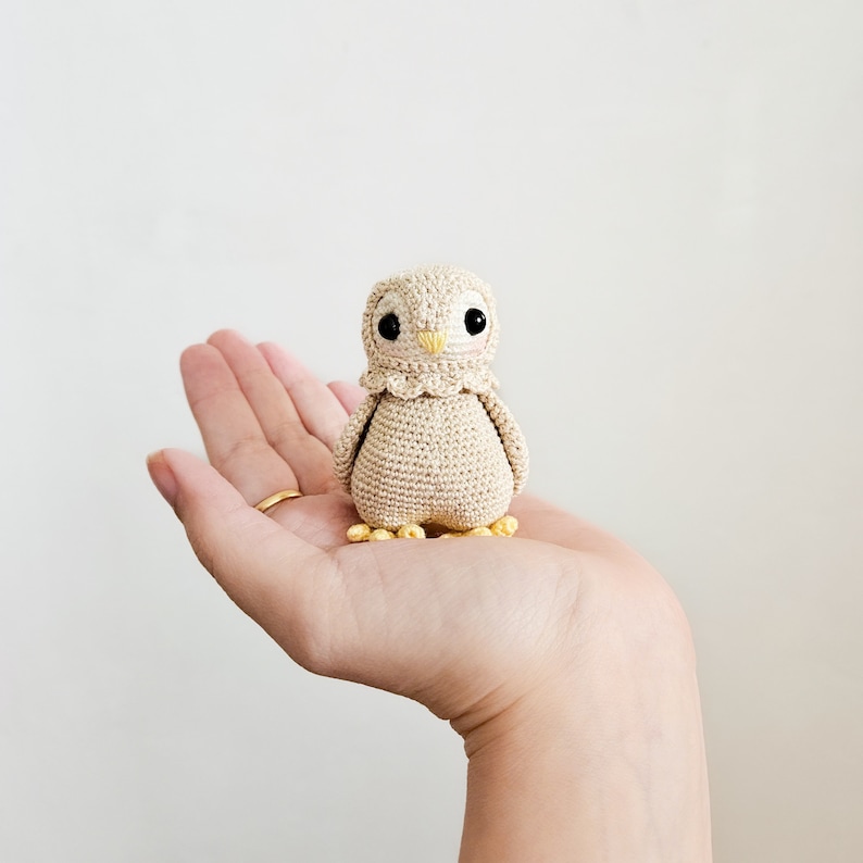 K&ouml;nnte beinhalten: Eine kleine geh&auml;kelte Eule aus beigefarbenem Garn mit gelben Akzenten. Die Eule sitzt auf einer Hand.