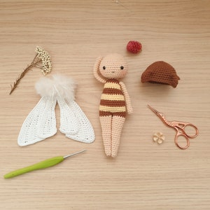 May include: A crocheted bee doll with brown and yellow stripes, white wings, and a brown hat. The doll is laying on a wooden surface with a crochet hook, scissors, and a small flower.