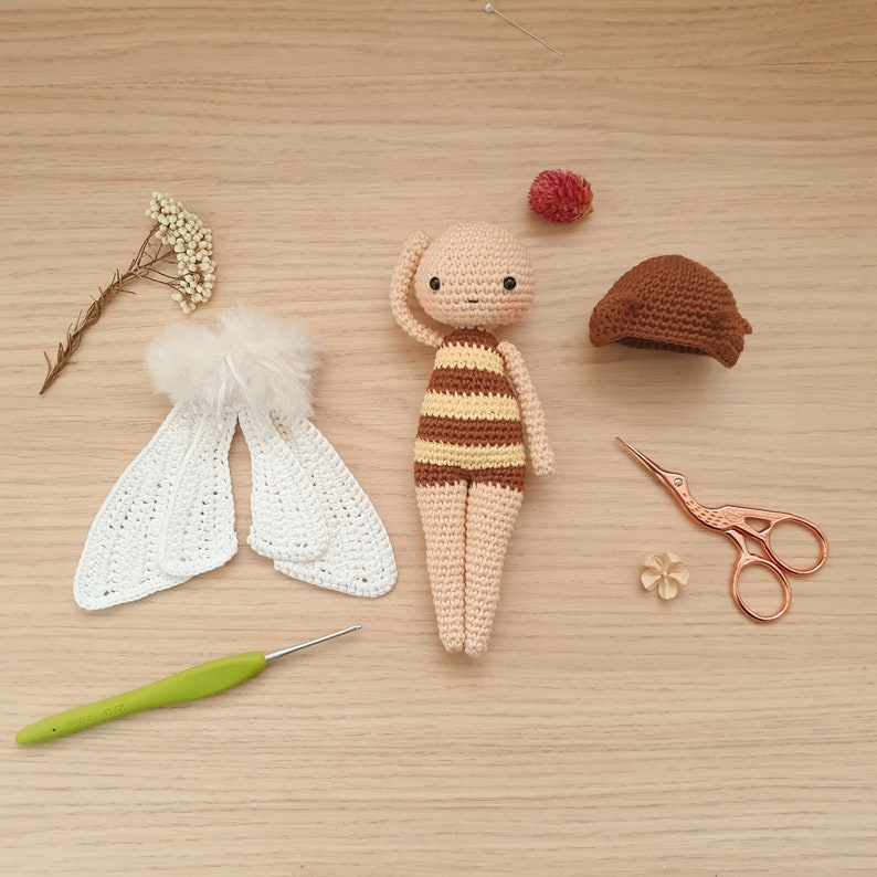 May include: A crocheted bee doll with brown and yellow stripes, white wings, and a brown hat. The doll is laying on a wooden surface with a crochet hook, scissors, and a small flower.