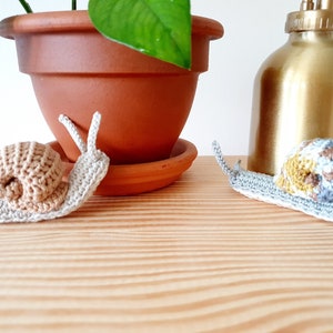 May include: Two crocheted snails, one beige and one with brown, yellow, and gray accents, on a wooden surface.