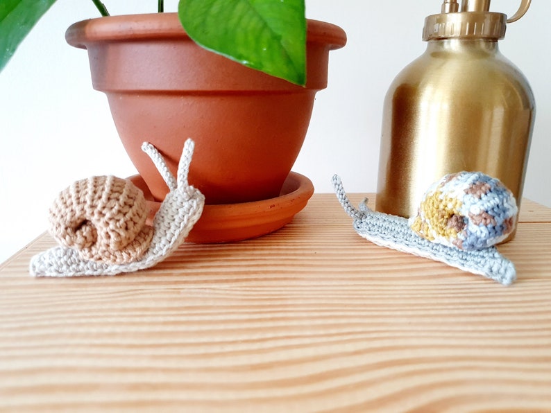 May include: Two crocheted snails, one beige and one with brown, yellow, and gray accents, on a wooden surface.