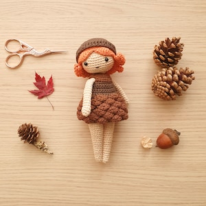 May include: A crocheted doll with brown hair, a brown hat, and a brown and cream dress. The doll is standing on a wooden surface with pine cones and a leaf.