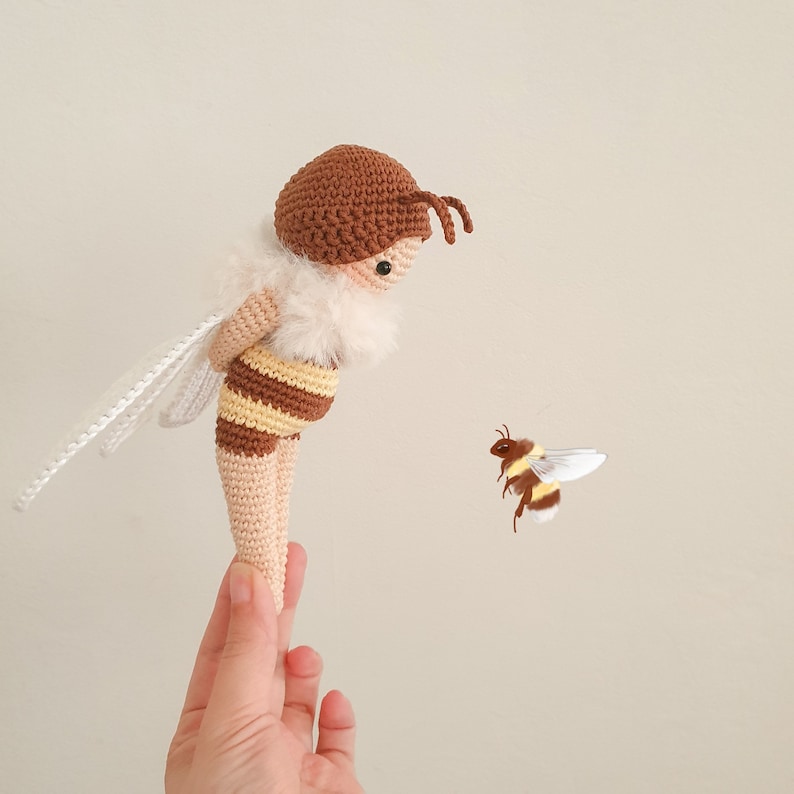 May include: A crocheted bee toy with brown and yellow stripes and white wings. The bee is standing on a hand, and a smaller crocheted bee is flying in the background.