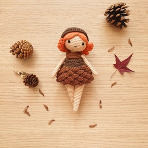 May include: A crocheted doll with brown hair, a brown dress, and a brown hat. The doll is sitting on a wooden surface with pine cones and leaves scattered around it.