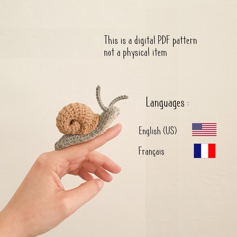 May include: A crocheted snail amigurumi toy in shades of brown and gray. The snail is sitting on a person's finger. The image includes the text "This is a digital PDF pattern not a physical item" and "Languages: English (US) Fran&ccedil;ais".