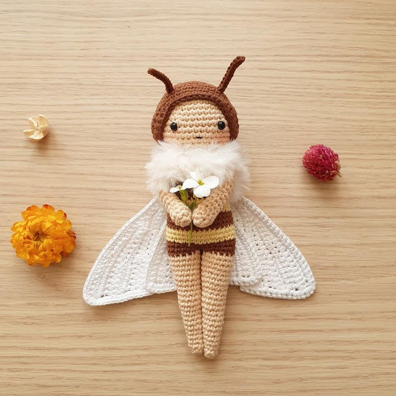 May include: A crocheted bee doll with brown and yellow stripes, white wings, and a fuzzy white chest. The doll is holding a bouquet of white flowers.