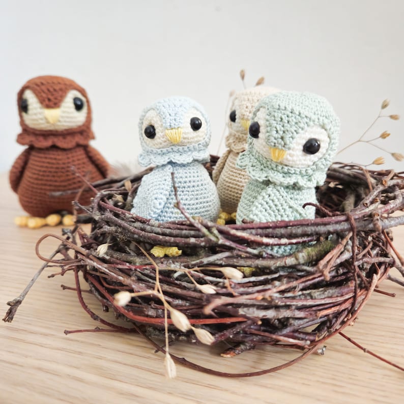 K&ouml;nnte beinhalten: Drei geh&auml;kelte Eulenfiguren in einem Nest aus Zweigen. Die Eulen sind hellblau, hellgr&uuml;n und braun.