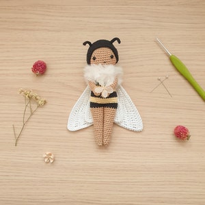 May include: A crocheted bee doll with black and yellow stripes, white wings, and a white flower in its hands. The doll is sitting on a wooden surface.