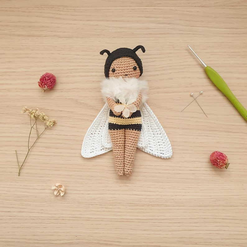 May include: A crocheted bee doll with black and yellow stripes, white wings, and a white flower in its hands. The doll is sitting on a wooden surface.