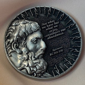 Puede incluir: Una moneda de plata con un retrato de Epicuro, un filósofo griego, en un lado. El otro lado presenta una cita de Epicuro: "No lo que tenemos, sino lo que disfrutamos, constituye nuestra abundancia." La moneda también incluye las fechas "BC 341-270".