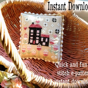 May include: A small, square, cross-stitch pillow featuring a house design with a red roof and black walls, surrounded by pink and white floral accents. The pillow is trimmed with white pom-poms and rests in a woven basket. Text reads "Instant Download".