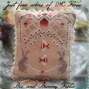 Op de afbeelding: Een beige kruissteek kussen met een bloemen- en konijnenontwerp. Het kussen heeft de tekst "Just four colors of DMC Floss" en "Bee and Bunny Frolic". Het ontwerp bevat roze, rode en bruine accenten, met een witte pompon-afwerking.