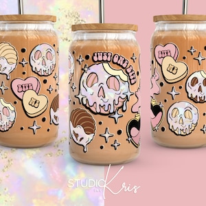 May include: Three glass tumblers with lids and straws, featuring a whimsical design of pink and white hearts, skulls, and apples with dripping icing. The tumblers are filled with a light brown liquid and have a brown background with gold stars.